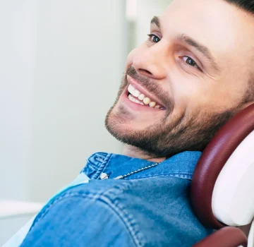 happy-man-with-hazel-eyes-dark-brown-hair-is-sitting-dental-chair-smiling-right-into-camera-because-he-is-satisfied-with-work-dentist